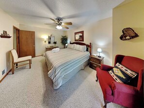 A hotel room with a bed, a chair, a desk, a nightstand, a lamp, a ceiling fan, a mirror, a door, and a wall-mounted shelf. - Pinon Park C11 | Cozy, Fireplace, Great Location (Ruidoso)