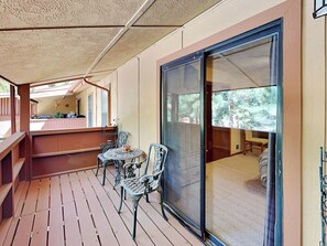 Una terraza de madera con puerta corredera de vidrio que da a un dormitorio. Hay dos sillas y una pequeña mesa en la terraza.