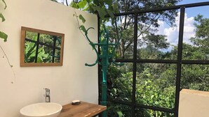 A bathroom with a wooden vanity, a white sink, a mirror, and a view of lush greenery through a large window. - Alban, Longo Mai - Cabaña Aqua - Via Namay Alto (Alban)