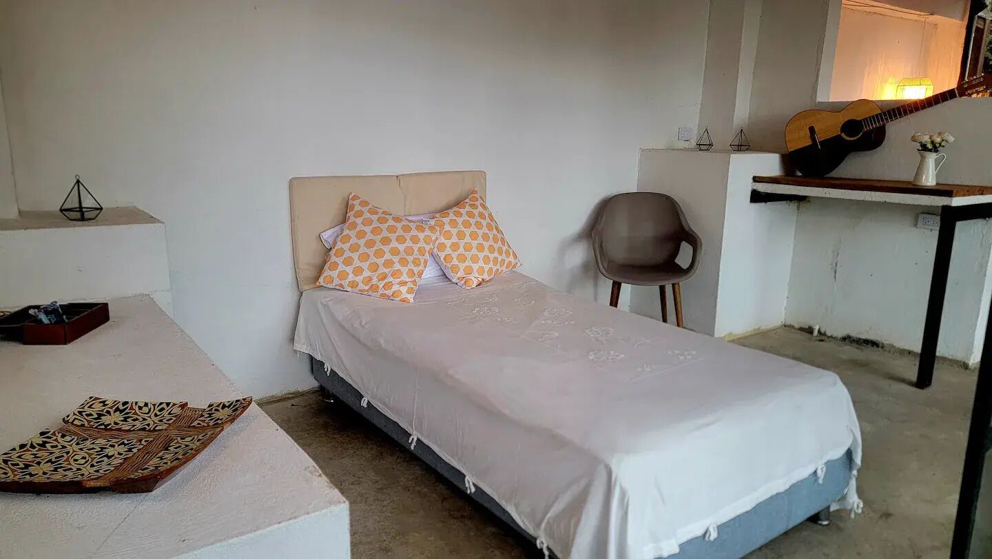 A single bed with patterned pillows, a chair, a guitar, and a small table with a vase.