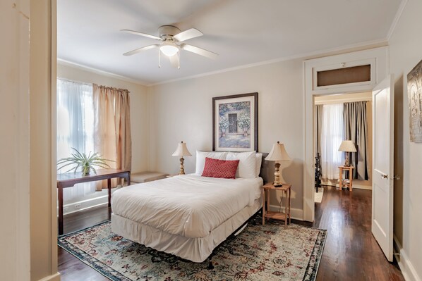 A bedroom with a bed, bedside tables, a ceiling fan, a window with curtains, and a door leading to another room. - Midtown Cottage Haute on Hannon (Mobile)