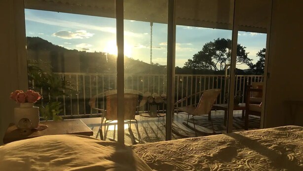 A bedroom with a large window overlooking a balcony. The room has a bed with a patterned bedspread, a small table with a vase of flowers, and a chair. The balcony has a table and chairs, and a view of trees and hills.