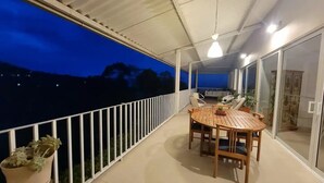 A balcony with a wooden table and chairs, potted plants, and a view of the night sky.