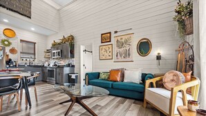 A spacious living area with a teal sofa, a glass coffee table, and a wooden chair. A kitchen with modern appliances is visible in the background.