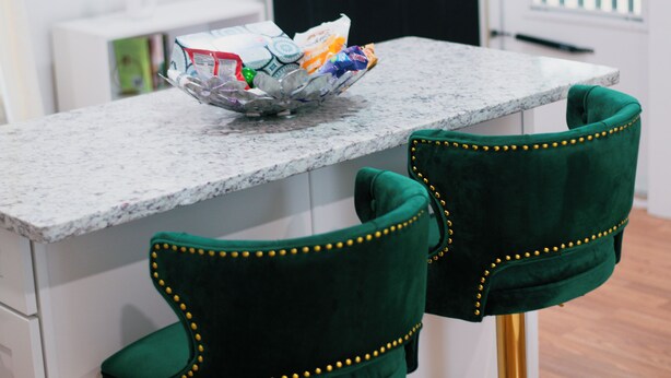 Una cocina con encimera de mármol, dos taburetes de bar tapizados en verde con detalles dorados y una puerta negra con ventanal.