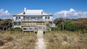 Une maison à deux étages située en bord de mer, dotée d’une terrasse en bois et d’un escalier menant à l’étage supérieur.