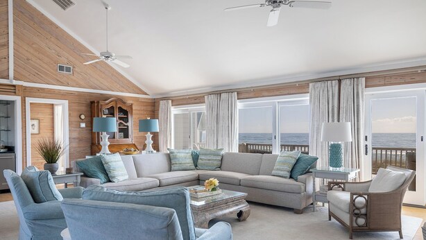 Un salon spacieux avec un grand canapé, des fauteuils et une table basse. On y trouve un ventilateur de plafond, une vue sur l’océan grâce à de grandes fenêtres et un mur en bois.