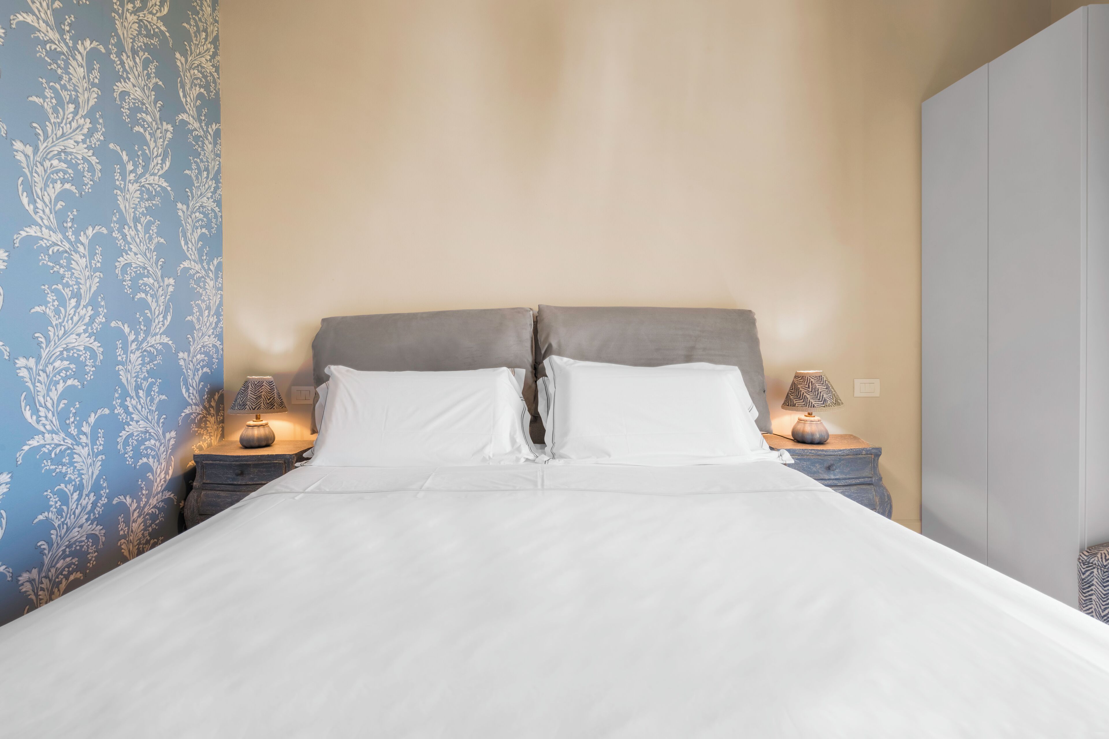 Una cama bien tendida con sábanas blancas en una habitación con papel tapiz azul estampado y una pared color beige.