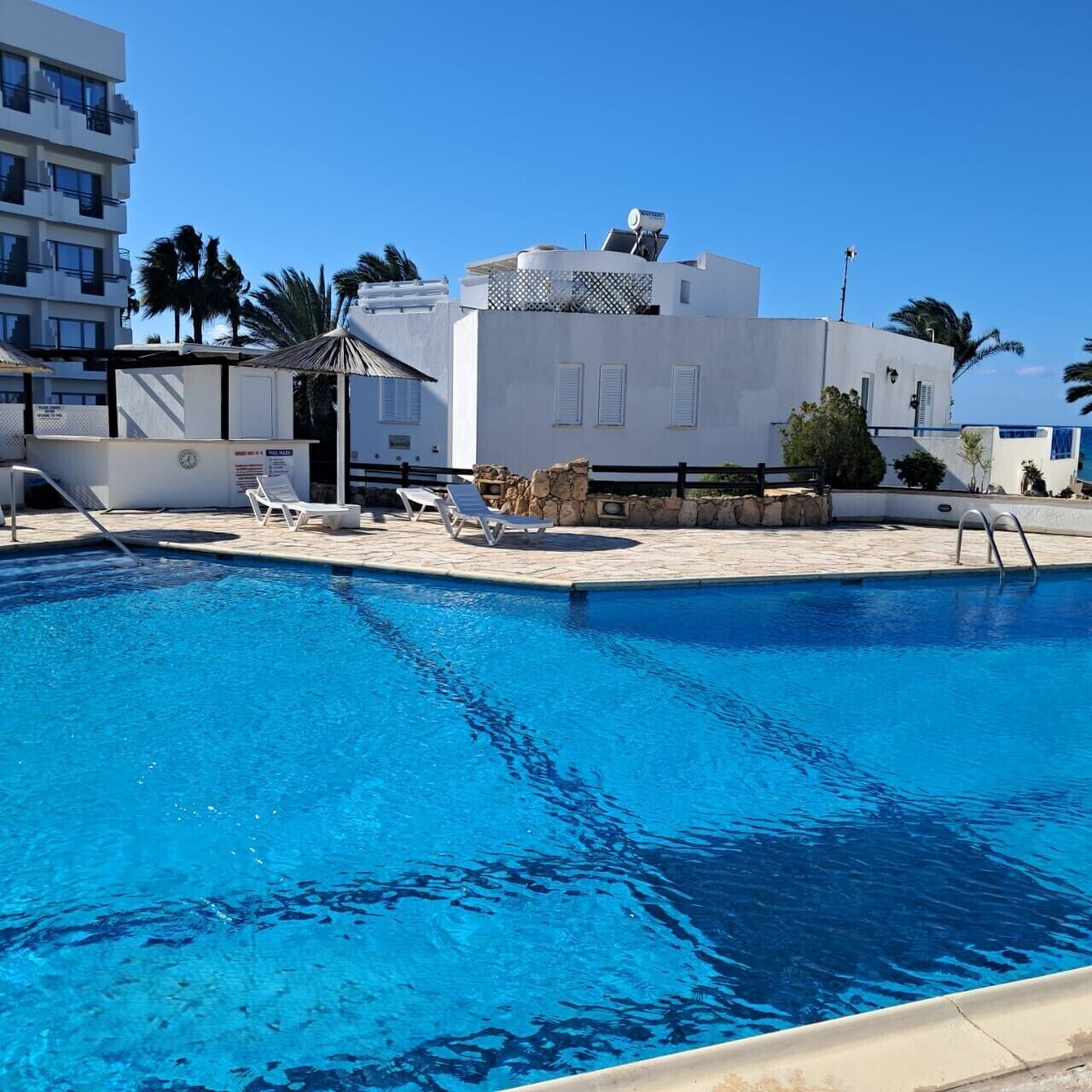 A swimming pool with lounge chairs and a stone wall, surrounded by a white building with a flat roof and a satellite dish.