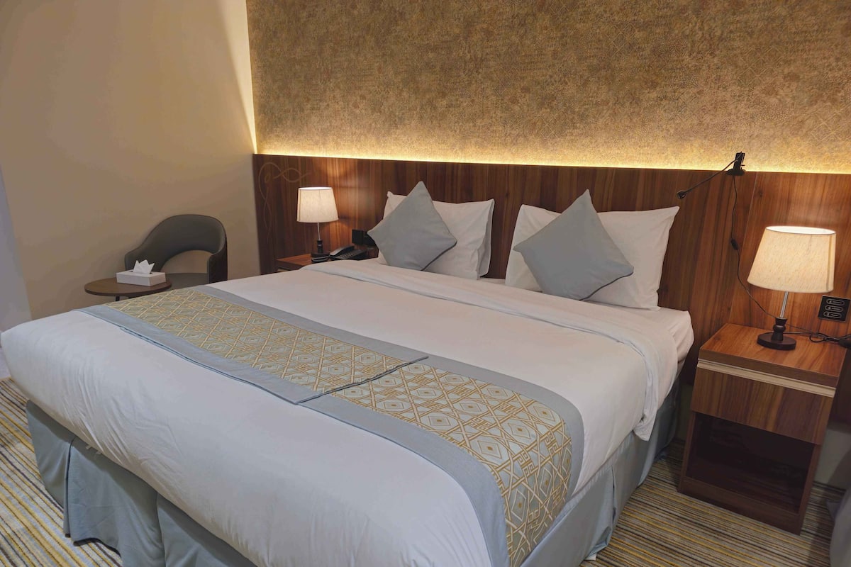 Una habitación de hotel con una cama grande, dos mesitas de noche con lámparas, una silla y un cabecero de madera.