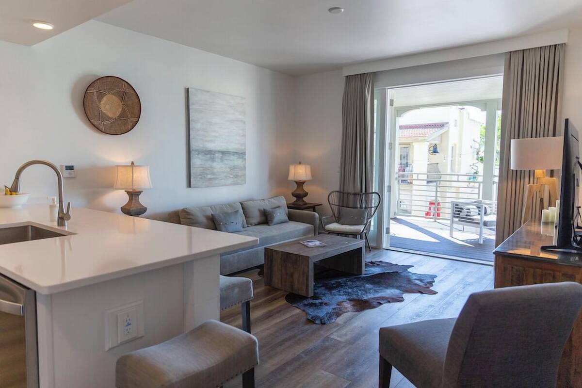 Een moderne woonkamer met een bank, salontafel en een eethoek. Er is een groot raam met gordijnen, een flatscreen-tv en een open haard.