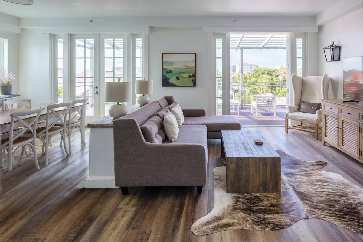 Een ruime woonkamer met een grijze bank, een houten bijzettafeltje en een groot vloerkleed.