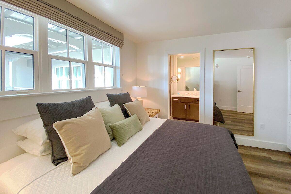 Een slaapkamer met een groot bed, een nachtkastje, een lamp, een spiegel en uitzicht op de badkamer door een open deur.