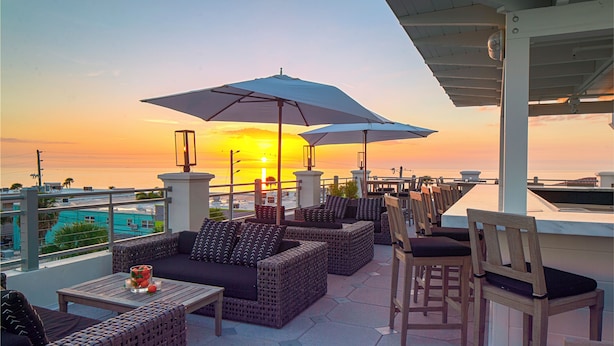 Una terraza con muebles de mimbre, sombrillas y vistas a la puesta de sol sobre el océano.