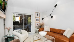 A modern living room with a brown leather sofa, white armchair, and a glass coffee table. There is a sliding glass door leading to a balcony, and a black floor lamp on the wall.