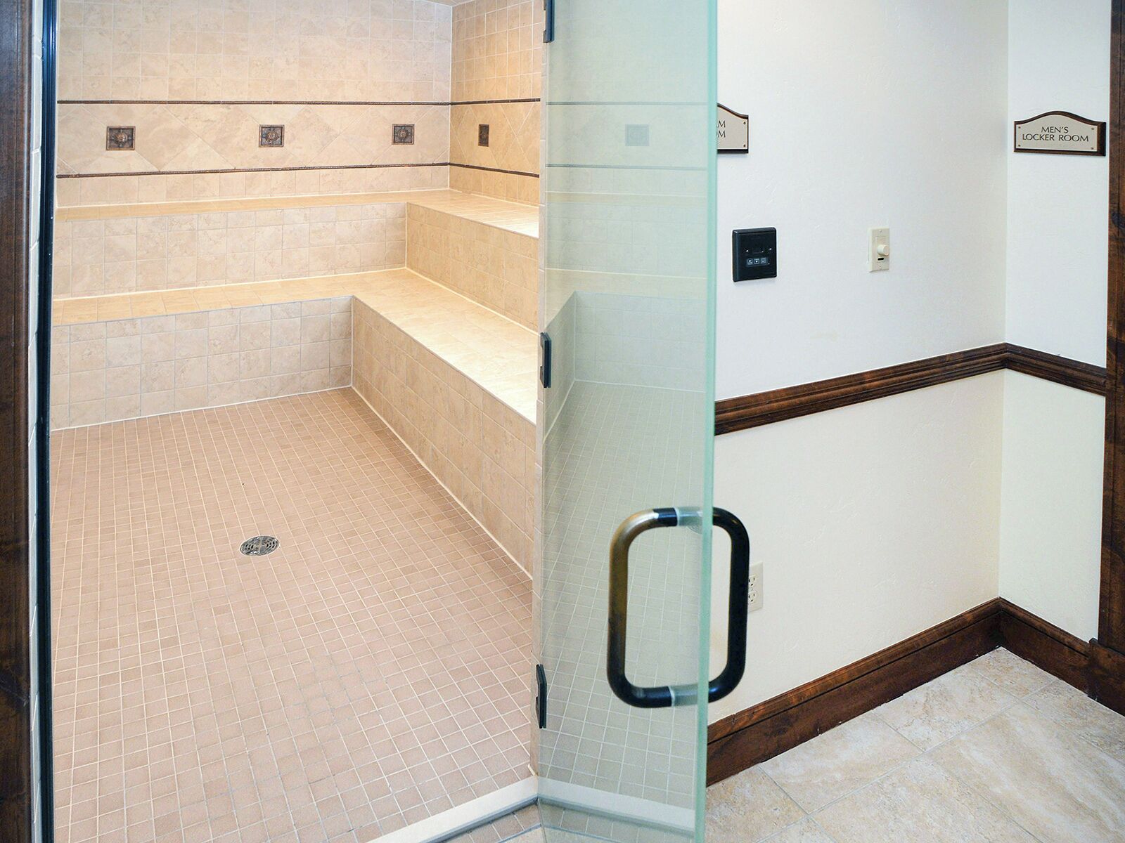 A men's locker room with tiled floors, a glass partition, and a wooden baseboard.