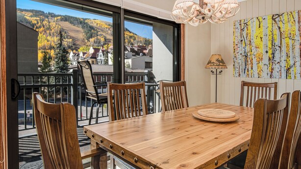Mesa de jantar de madeira com seis cadeiras, um candelabro e vista para árvores e edifícios outonais através de grandes janelas.