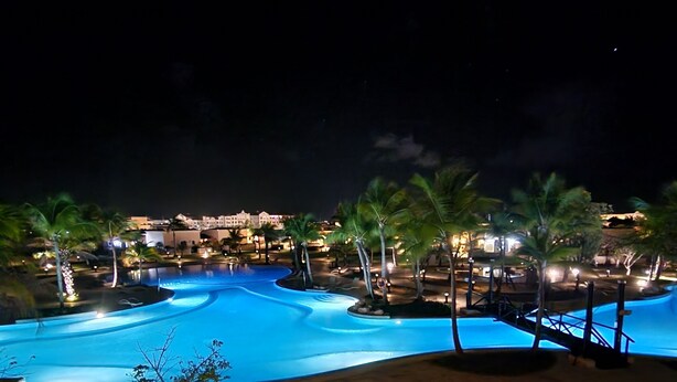 A nighttime view of a resort with a large swimming pool, palm trees, and well-lit pathways.