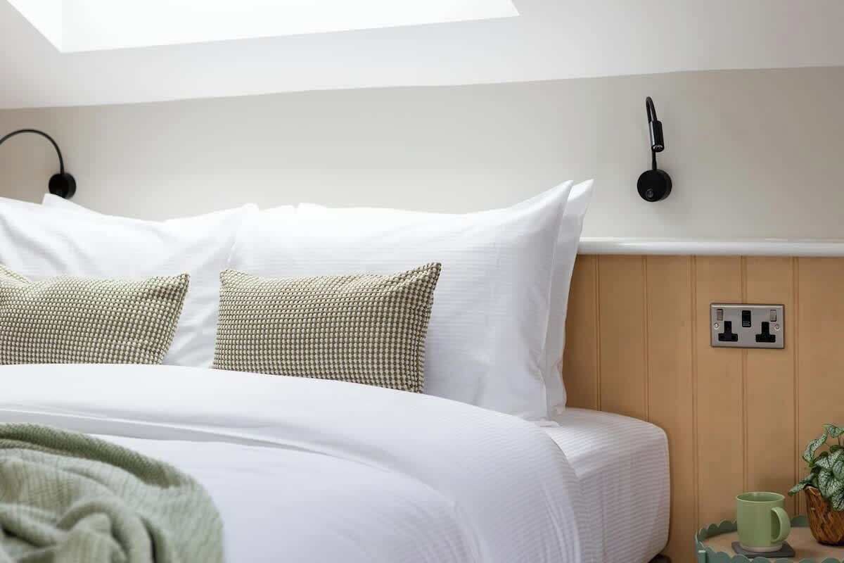 A neatly made bed with white and patterned pillows, a bedside lamp, and a power outlet on the wall.