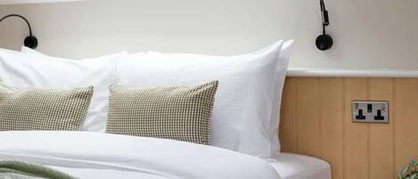 A neatly made bed with white and patterned pillows, a bedside lamp, and a power outlet on the wall.