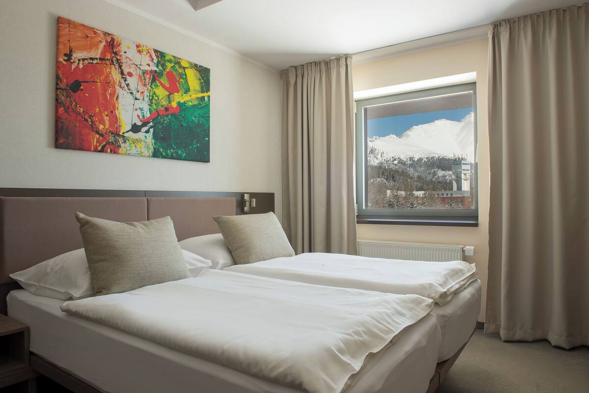 A hotel room with two beds, a large abstract painting on the wall, and a window with a view of snow-capped mountains.