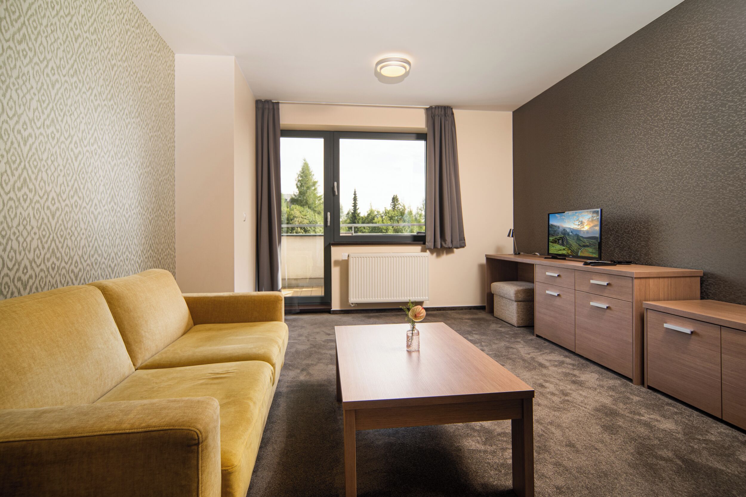 Una habitación de hotel moderna con sofá, una mesa de madera, televisión y vistas a zonas verdes.