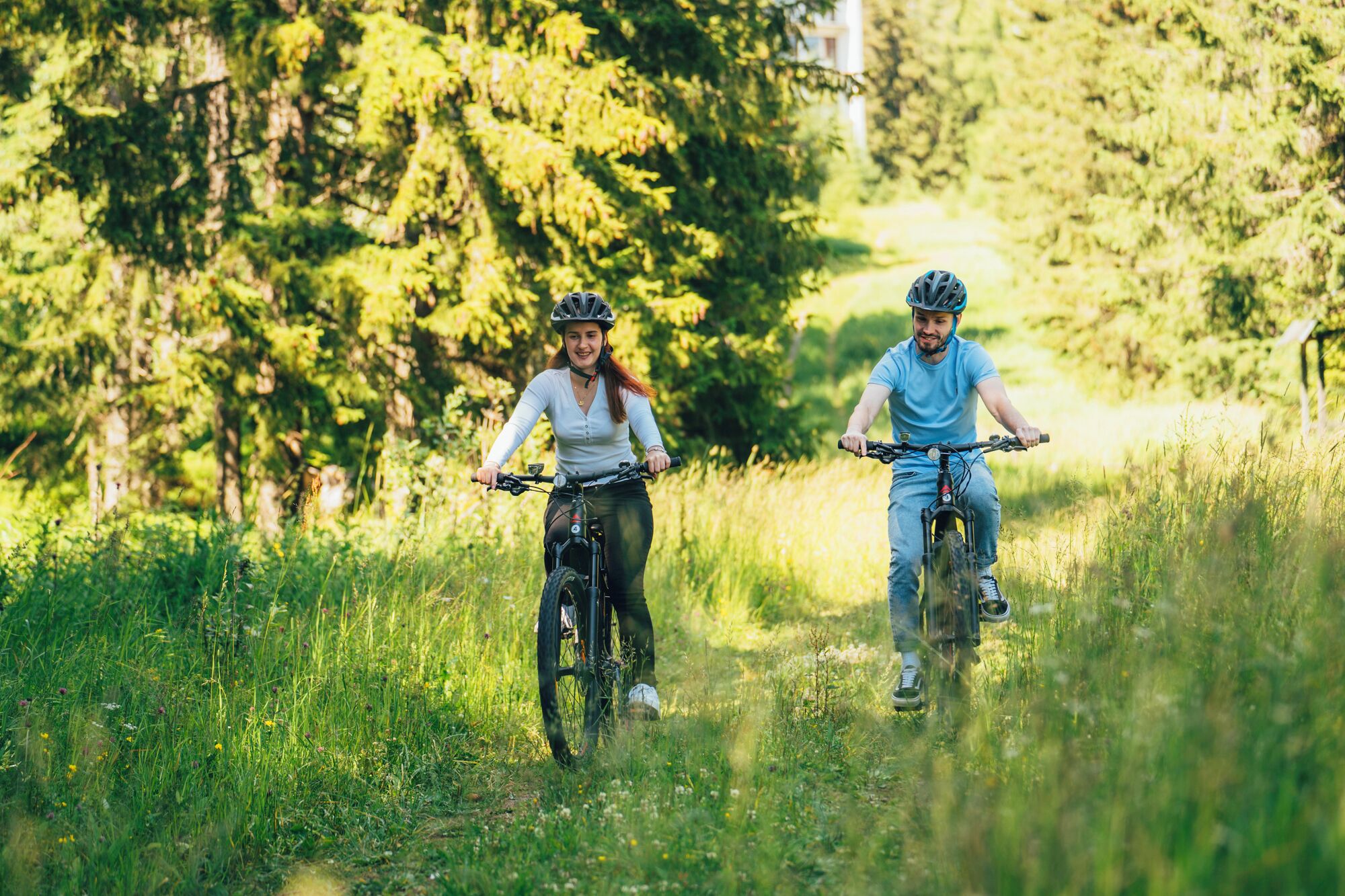Two people riding bicycles on a grassy path surrounded by trees.