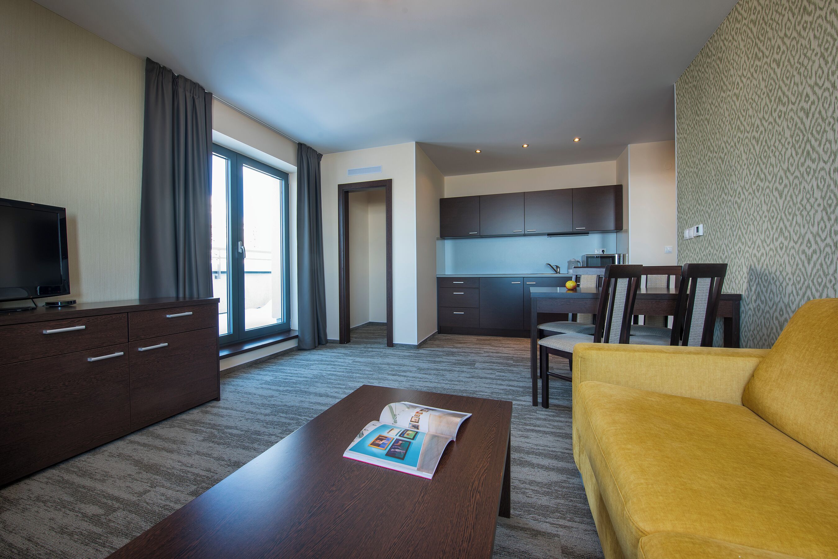 Una habitación de hotel moderna con televisor de pantalla plana, un comedor con mesa y sillas, una zona de cocina y un amplio ventanal con cortinas.