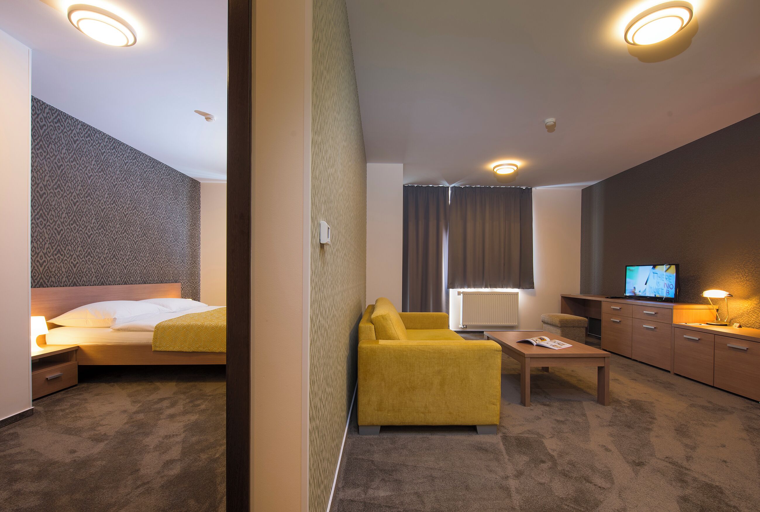 A hotel room with a bed, a yellow armchair, a coffee table, a TV, and a desk with drawers.