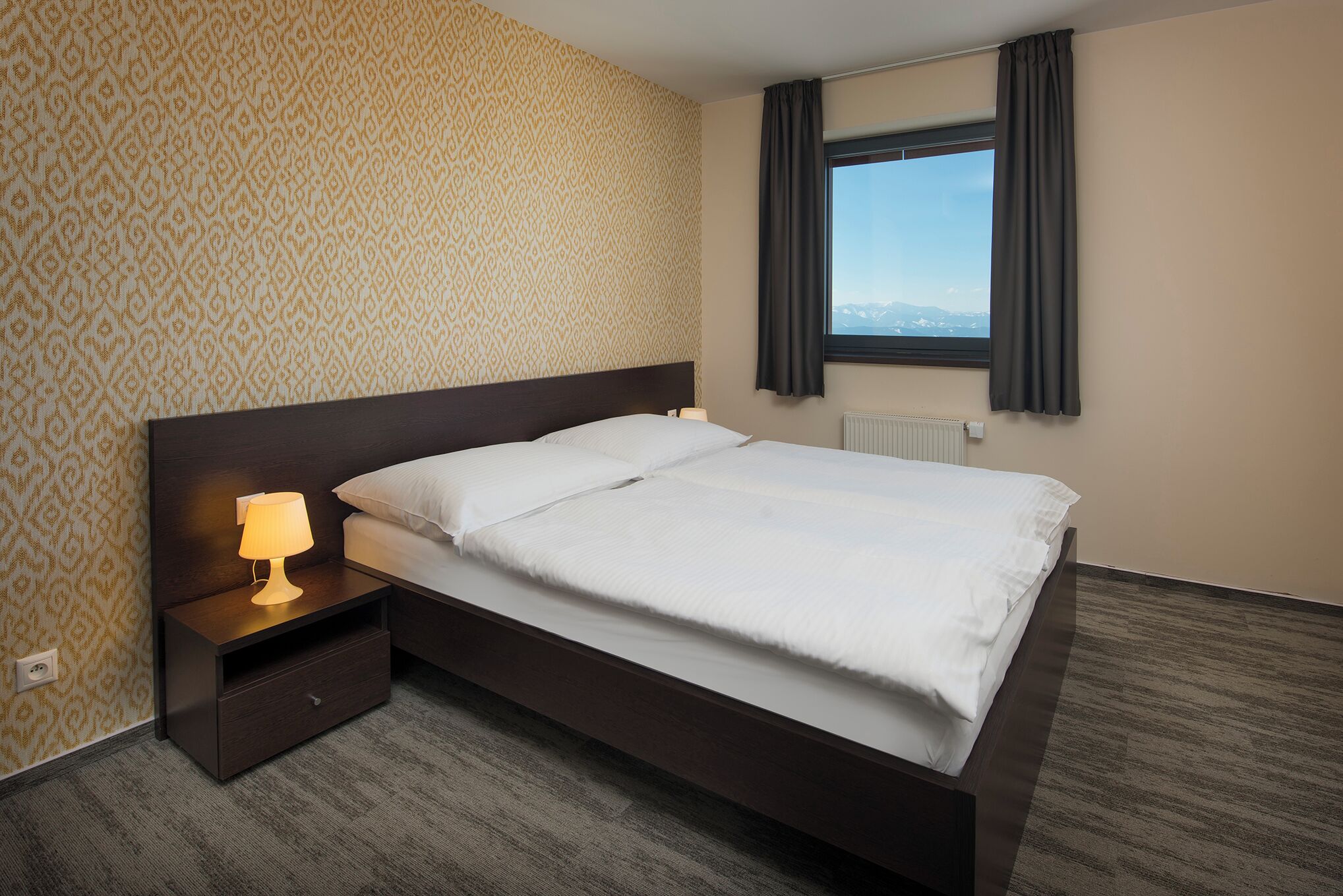 A hotel room with a large bed, a nightstand with a lamp, patterned wallpaper, a window with curtains, and a view of the mountains.