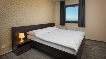 A hotel room with a large bed, a nightstand with a lamp, patterned wallpaper, a window with curtains, and a view of the mountains.
