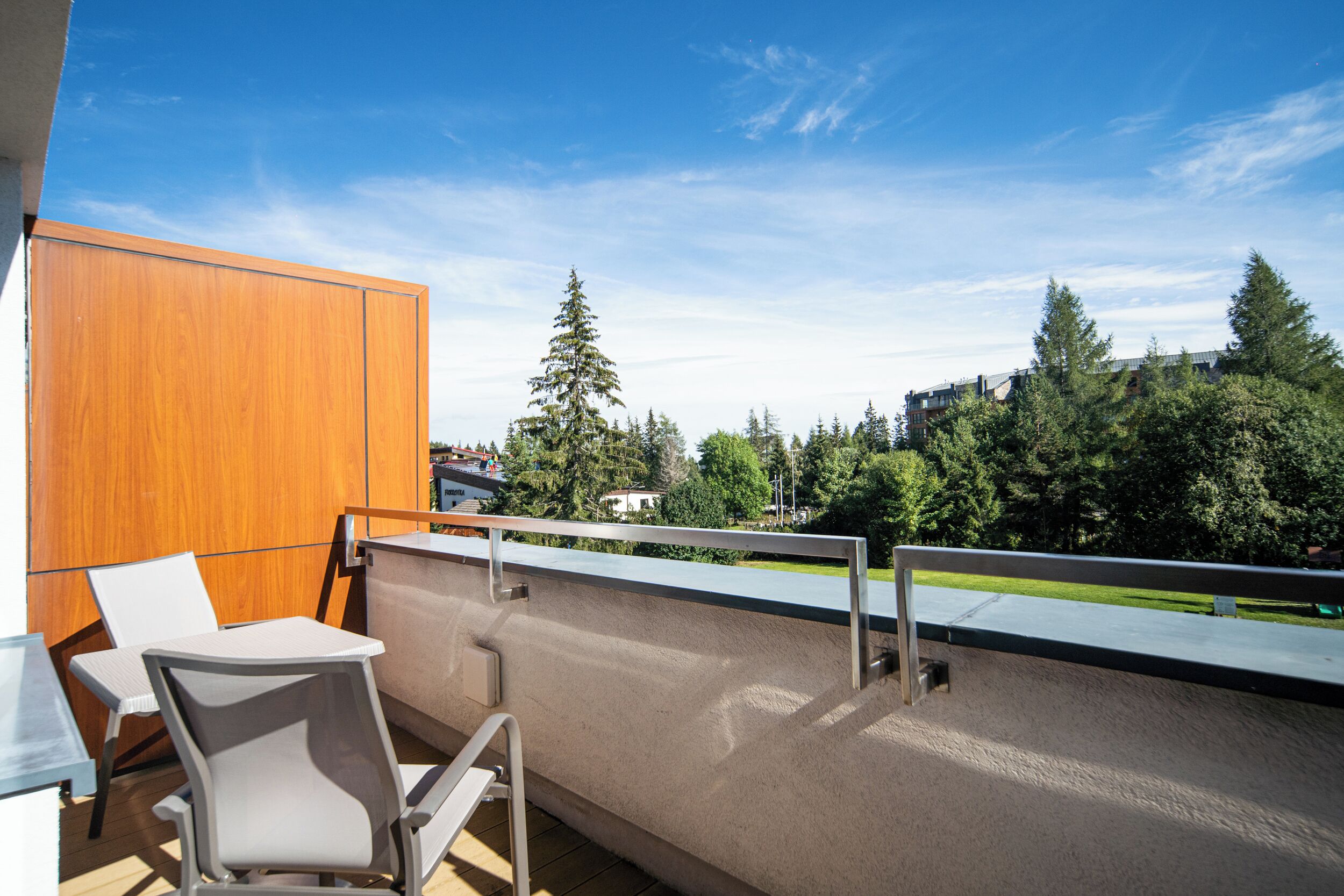 A balcony with a view of trees and buildings, a wooden panel, and a table with chairs.