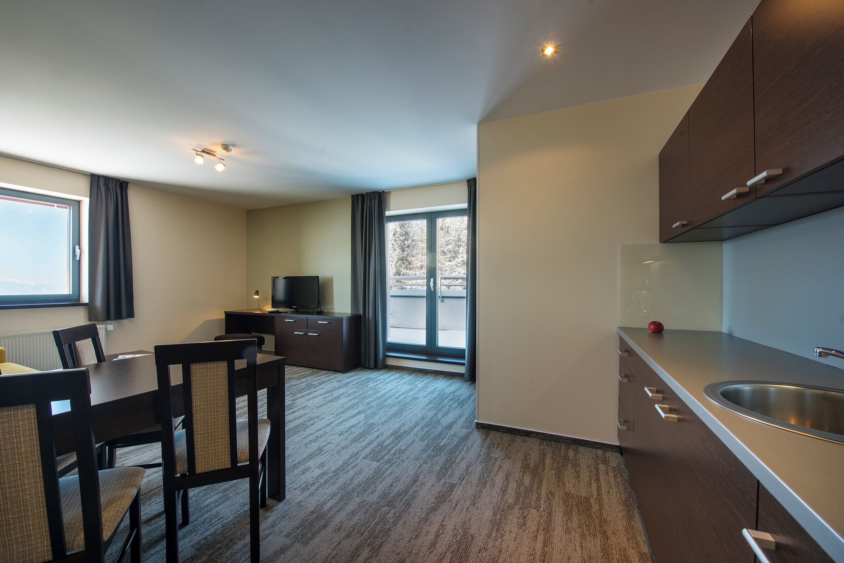 A modern kitchen with a dining area, large windows, and a flat-screen TV.