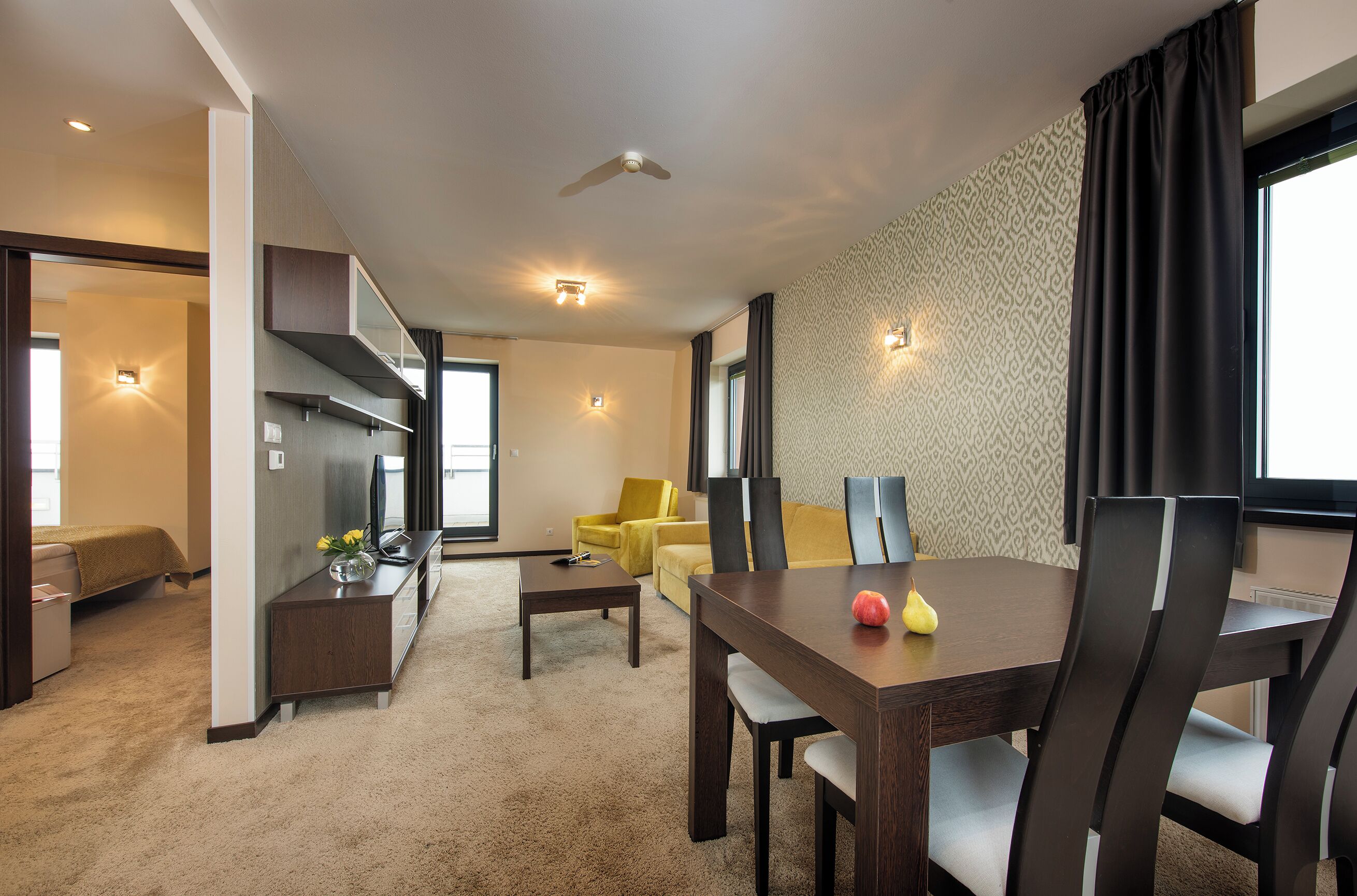 A modern hotel room with a dining area, a sofa, a TV, and a ceiling fan.