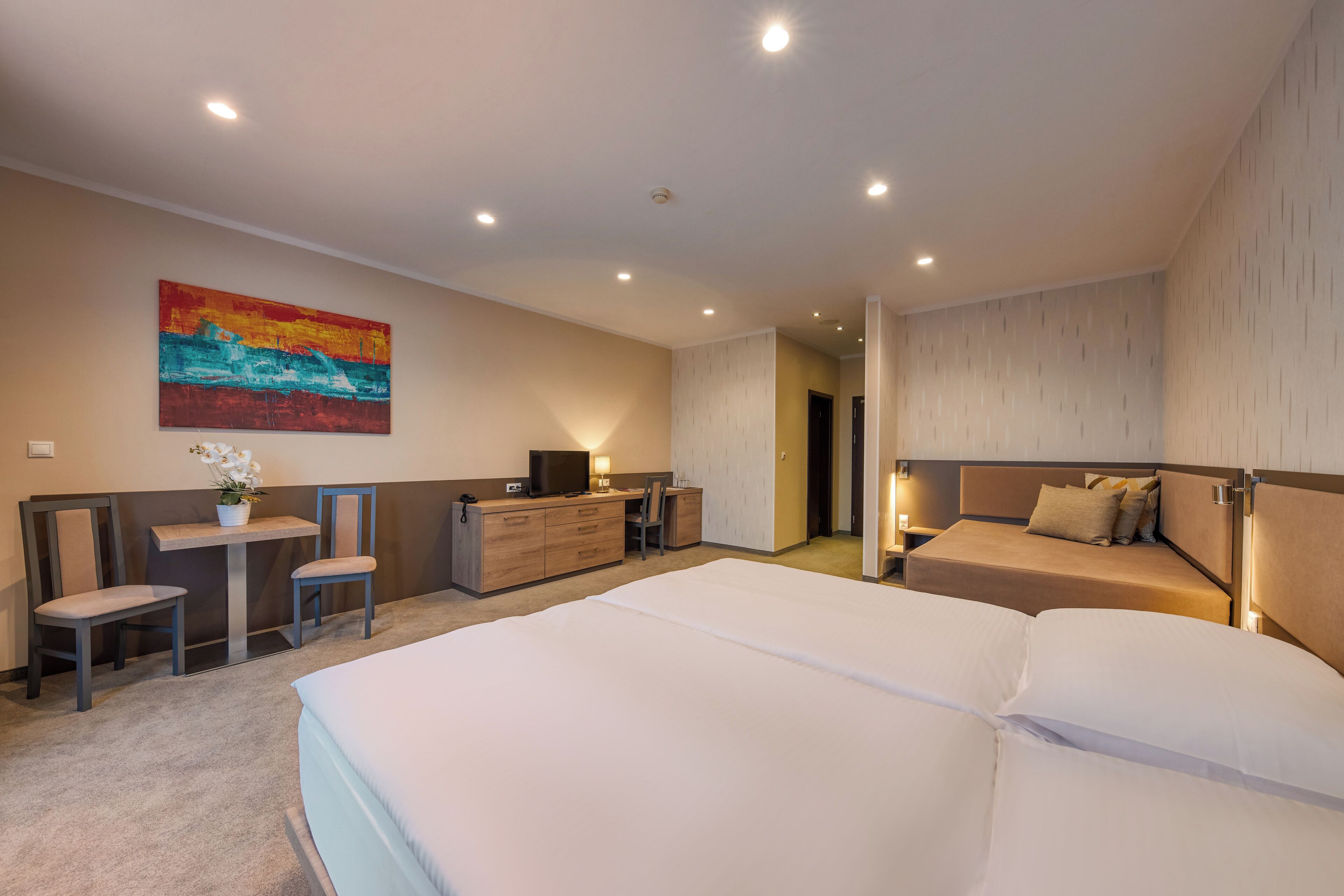 A hotel room with a large bed, a desk with a chair, a small table with a plant, and a painting on the wall.