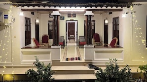 A hotel named Coconut Creek with a sign stating "Home Stay is Here to Stay." It features a central doorway flanked by two red chairs and a bench, with string lights and potted plants in the foreground. - Coconut Creek Homestay (Kottayam)