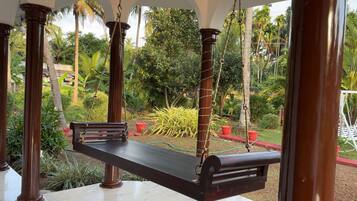 Una hamaca de madera colgada entre columnas en un jardín exuberante, con palmeras y plantas en macetas.