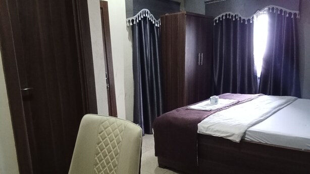 A hotel room with a bed, a chair, a wardrobe, and curtains. - Dutse Suites and Luxury (Abuja)
