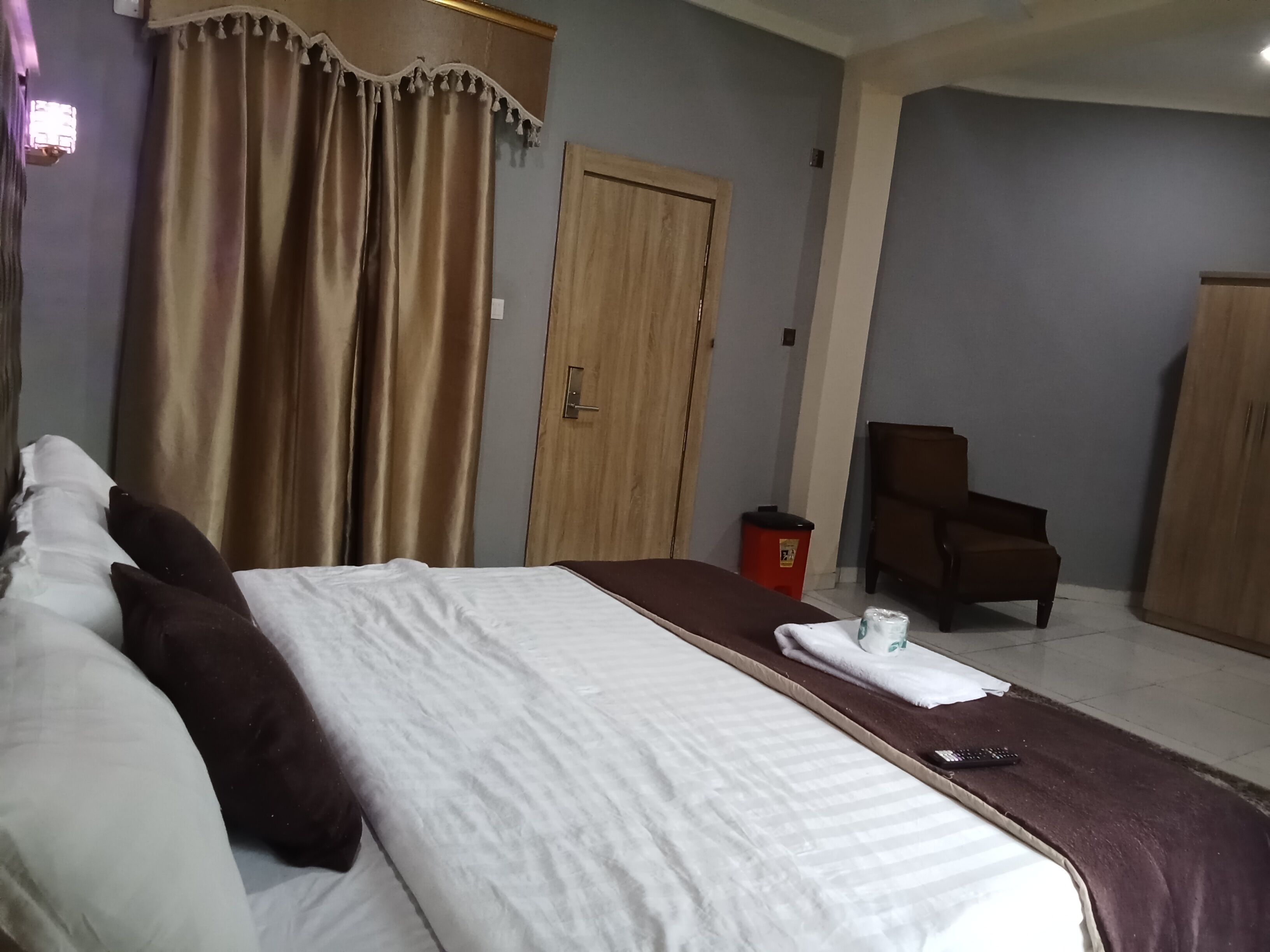 A hotel room with a large bed, a wooden wardrobe, a chair, and a small trash bin.