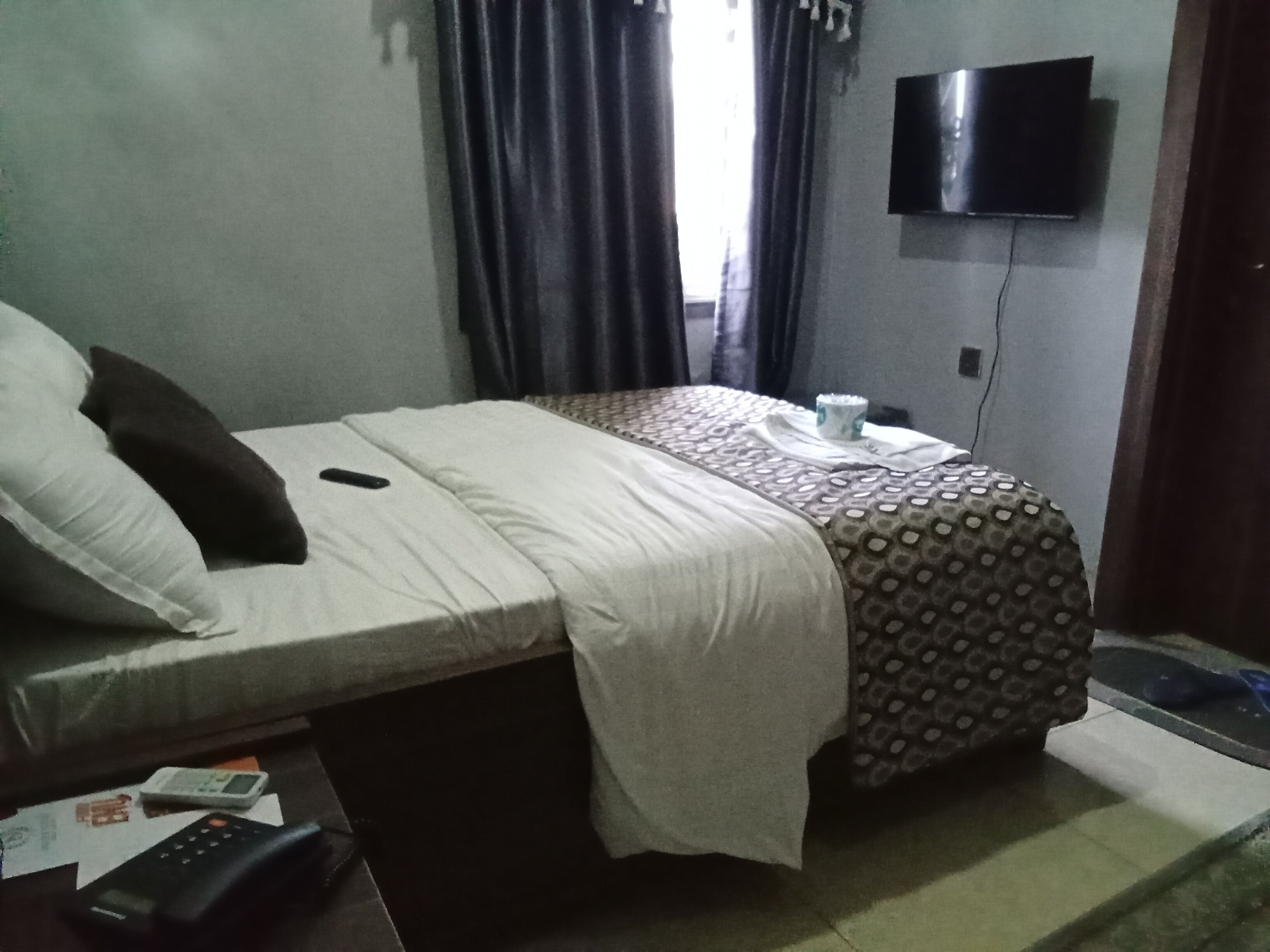 A hotel room with a bed, a television mounted on the wall, a small table with a remote control, and a window with curtains.