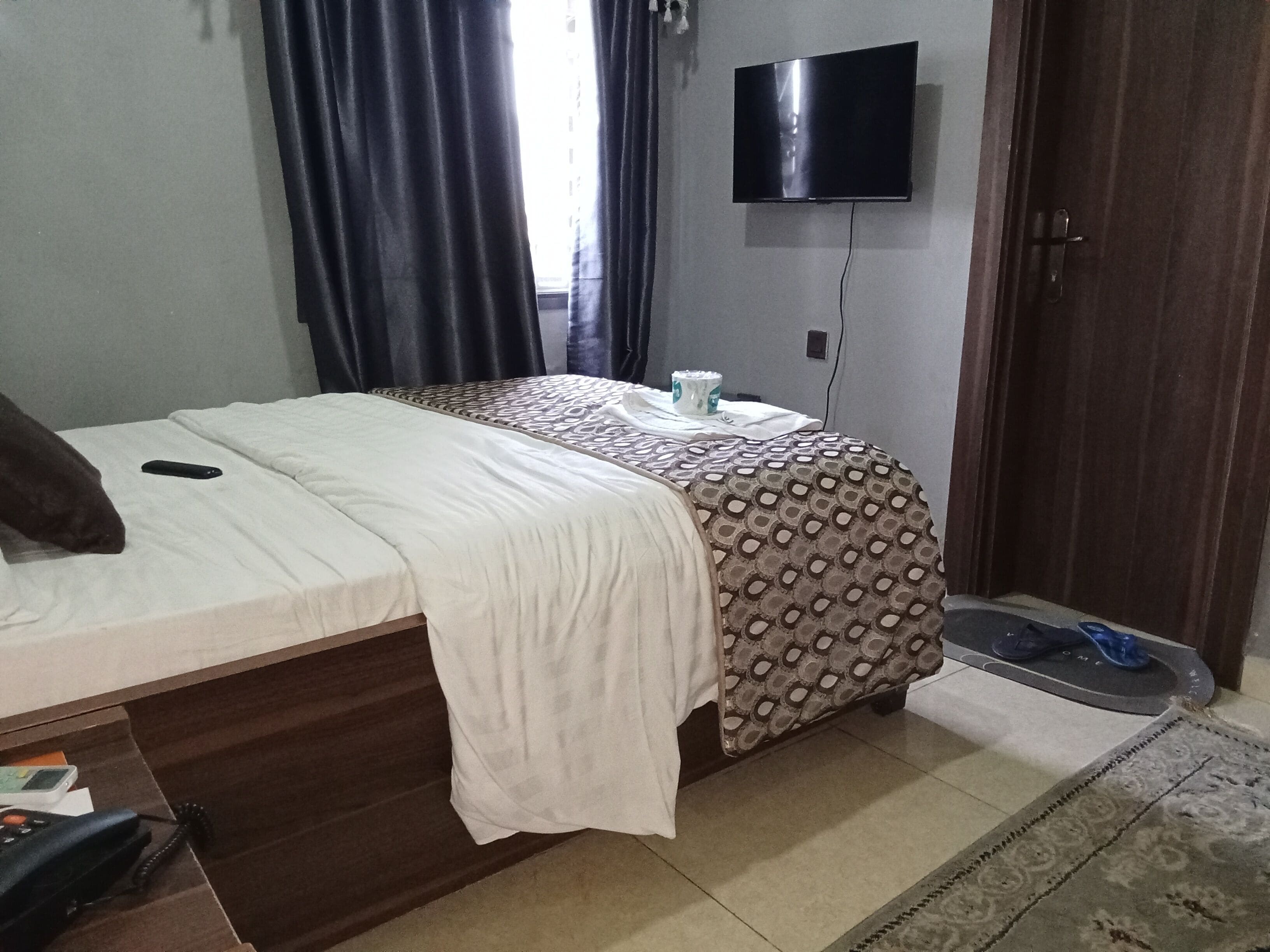 Una camera da letto con un letto, una TV a muro, un comodino con una tazza e una porta con un tappetino.
