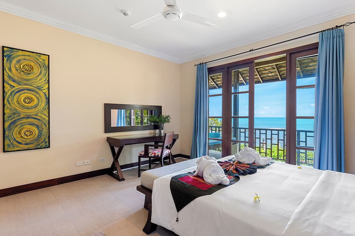A hotel room with a bed, a desk, a chair, a large window with a view of the sea, and a painting on the wall.