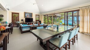 Een ruime woonkamer met een grote glazen tafel, blauwe zachte zitting, een televisie en uitzicht op de oceaan.
