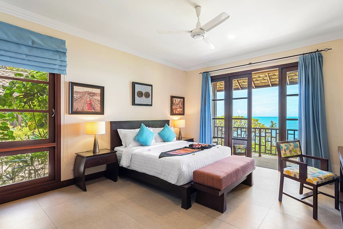 A hotel room with a bed, bedside tables, a desk, a chair, and a balcony with a view of the sea.