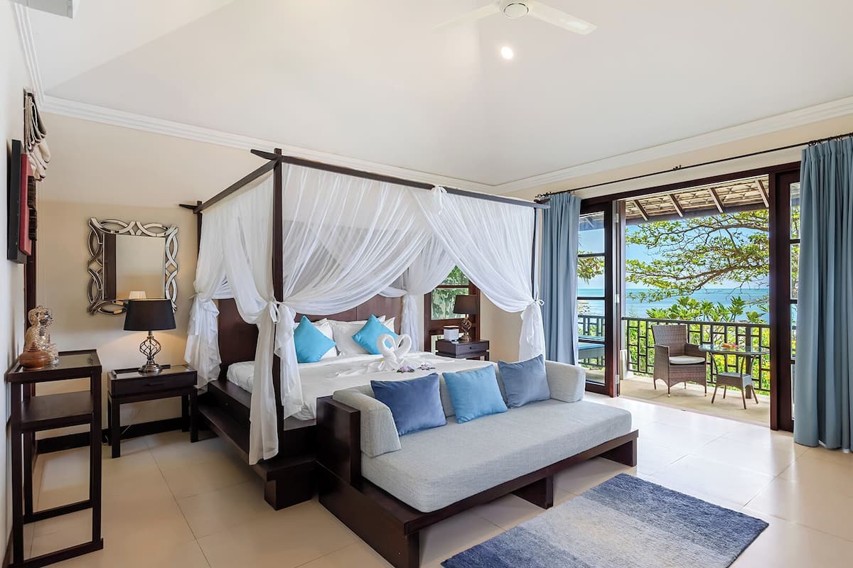 A bedroom with a four-poster bed, a sofa, a balcony with a view of the sea, and a large mirror on the wall.