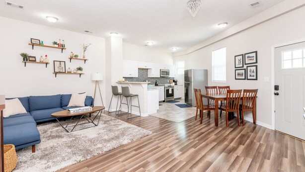 Un vaste espace de vie comprenant un canapé bleu, une table basse et une table à manger avec des chaises. On aperçoit la cuisine à l’arrière-plan, avec des meubles blancs et un four à micro-ondes.