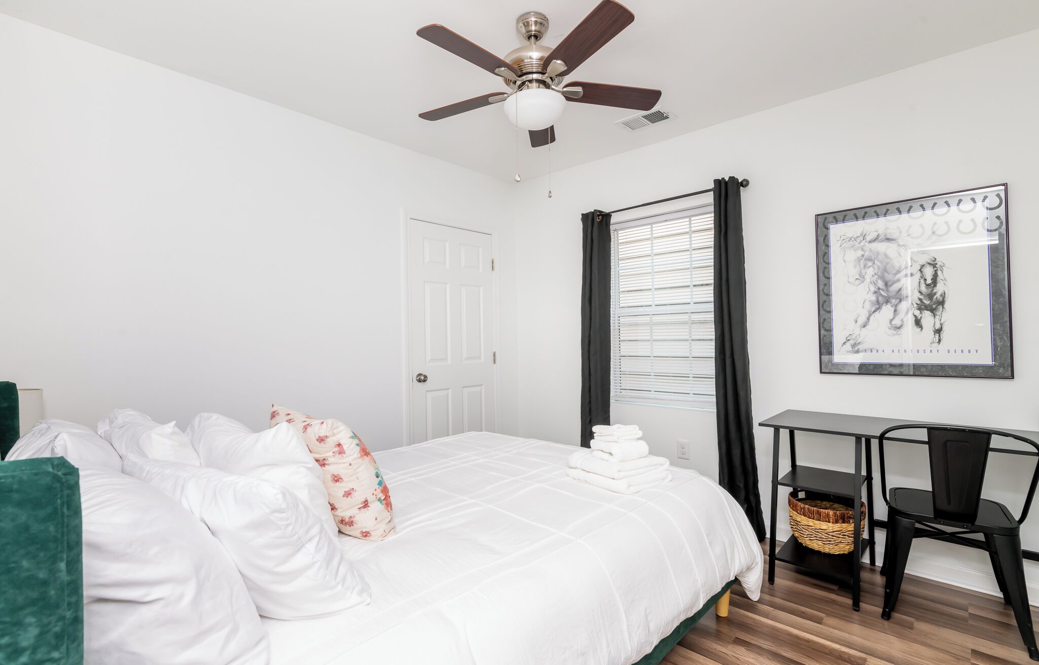Une chambre à coucher comprenant un lit, un bureau, une chaise, un ventilateur de plafond et une fenêtre avec des rideaux.
