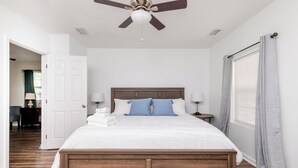 Un lit bien fait, avec une tête de lit en bois, une literie blanche et deux oreillers bleus. Un ventilateur de plafond est installé au-dessus du lit, et une fenêtre avec des rideaux se trouve sur le côté droit.