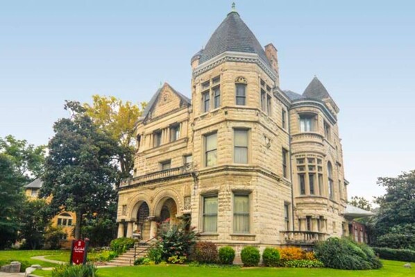 A large, historic stone building with multiple towers and arched windows, surrounded by well-maintained gardens.