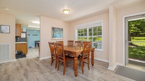 A dining area with a wooden table and chairs, a kitchen in the background, and a view of a green lawn through a window. - New! Lakefront, Great Location, Pet Friendly (Henrico)